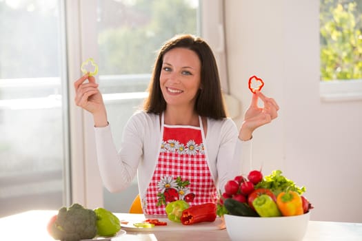 Happy young woman sitting at table with vegetables in bowl and holding pepper slices in hands