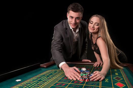 Young beautiful couple takes their winnings at the roulette table at the casino, on a black background. A man in a suit with a woman in a black dress