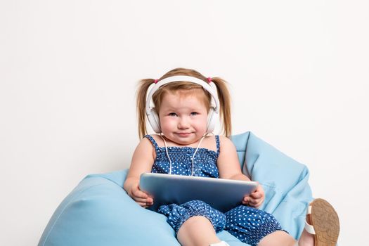 Cute little girl in headphones listening to music using a tablet and smiling while sitting on blue big bag. On white background. A child looks at the camera