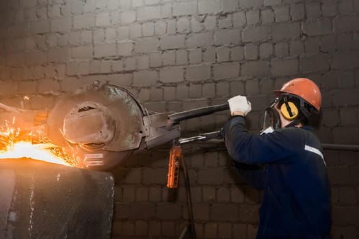 A male worker in a protective helmet, respirator, overalls manages heavy grinding equipment for cast iron concrete tubing with flying sparks in the workshop of an industrial plant.