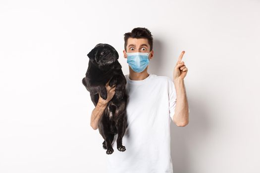 Covid-19, animals and quarantine concept. Young man in medical mask holding cute black pug, dog looking right and owner pointing at logo, white background.