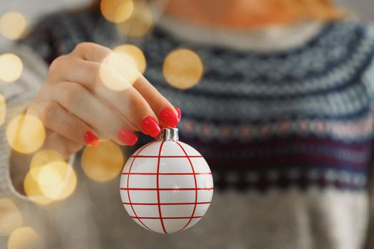 Hand of a woman in festive sweater holding Christmas bauble with blurred lights close up