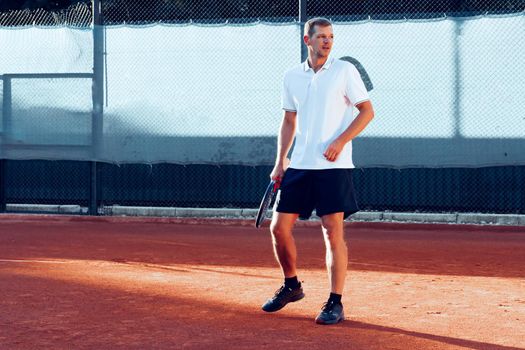 Young man plays tennis outdoors on clay tennis court in the morning