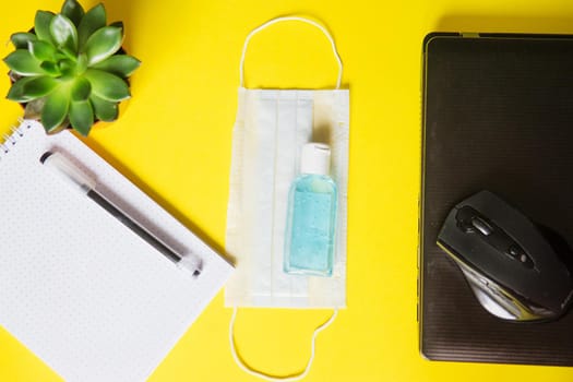 Top view of a desktop with notebooks, pens, and a laptop. Work from home using an online device, protection from coronavirus during the quarantine period. Protective masks and antiseptic on a yellow background.