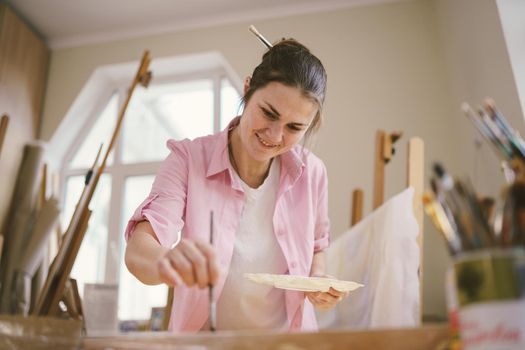 Cute woman paints on canvas in an art workshop. Artist creating picture. Art school or studio. Work with paints, brushes and easel. Hobby and leisure concept. Woman painter at workspace.