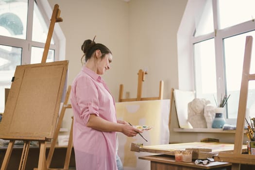 Cute woman paints on canvas in an art workshop. Artist creating picture. Art school or studio. Work with paints, brushes and easel. Hobby and leisure concept. Woman painter at workspace.