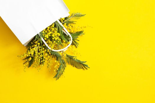 A bouquet of mimosa in a white mock up gift bag. Spring shopping, gifts and promotions for International Women's Day. Yellow background, copyspace.