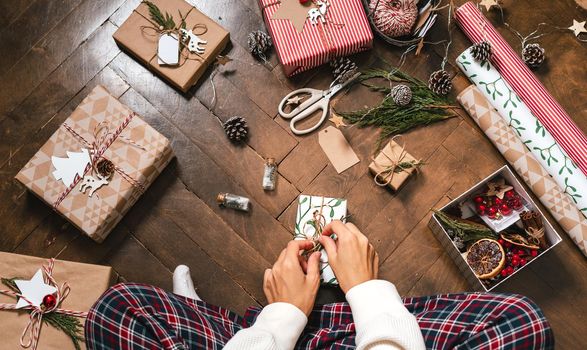 Woman s hands wrapping Christmas gift boxes, close up. Cropped female sit and preparing natural eco presents on floor with decor elements and items. Merry Christmas or New year DIY packing Concept.