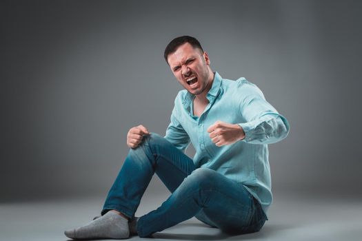 The man is sitting on the floor, isolated on gray background. Man showing different emotions. A man is dressed in blue jeans and a shirt