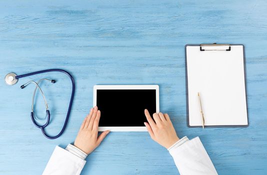 Top view of doctor hands with tablet computer. Therapist sitting at blue desk with stethoscope and clipboard. Examination and consultation in clinic. Medical application and online healthcare services
