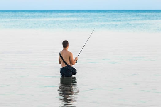 A young guy without a shirt stands knee-deep in the water of the sea in the evening and catches fish with a line.