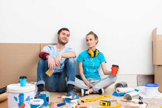 Happy boy and girl drinking coffee on floor. Home remodeling after moving. Cardboard boxes, construction tools and materials for building on floor. Couple having fun in time of house renovation.