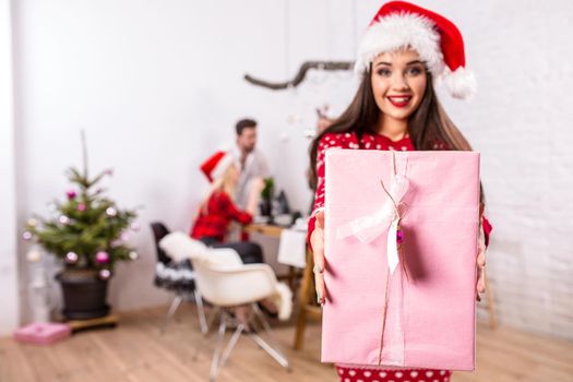 Portrait of a young woman with a pink giftbox on the foreground. Beautiful brunette in a Santa hat, red costume with deers. Christmas party at home.