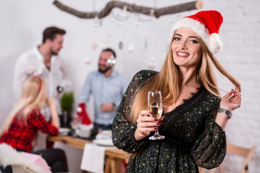 Portrait of a young woman with a glass of champagne on the foreground. Beautiful blonde in a Santa hat and black dress. Christmas party at home.