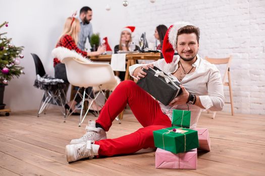 Shot of happy friends enjoying holidays. Focus on the man in the foreground in a red Christmas hat. A man with a gift boxes in his hands in a white room