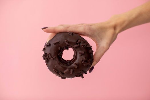 Crop female hand holding sweet chocolate donut isolated on pink background