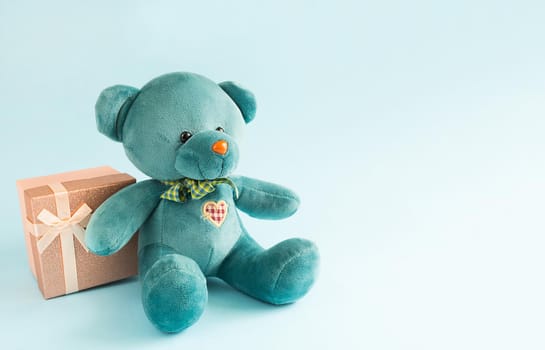 Turquoise soft teddy bear with an embroidered heart holds a gift box and a bow on a blue background. Children's toy. Love, a gift to holiday, a declaration of love, Valentine's day. Copy space