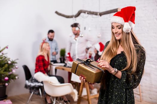 Portrait of a young woman with a gold giftbox on the foreground. Beautiful blonde in a Santa hat and black dress. Christmas party at home.