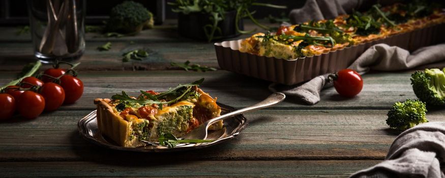 Slice of delicious homemade quiche with cherry tomatoes and broccoli on metal plate. Dark photo of healthy vegetarian food.