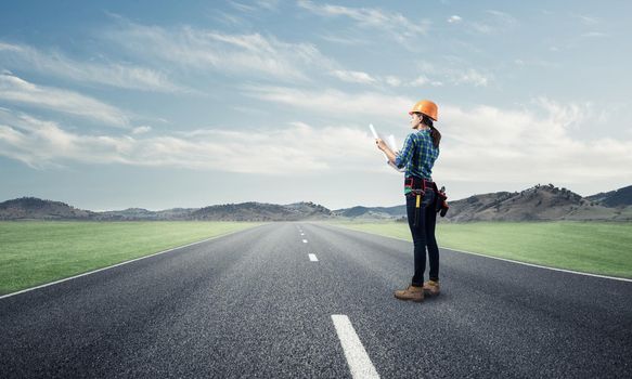 Young woman in safety helmet standing on asphalt highway with blueprint. Back view architect in workwear checking construction on nature background. Road infrastructure reconstruction and development