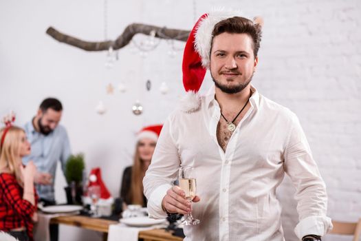 Shot of happy friends enjoying holidays. Focus on the man in the foreground in a red Christmas hat. Man with a glass of champagne in a white room