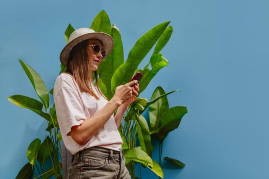 Pretty young woman in stylish hat and sunglasses uses smartphone while standing near green plant on blue background