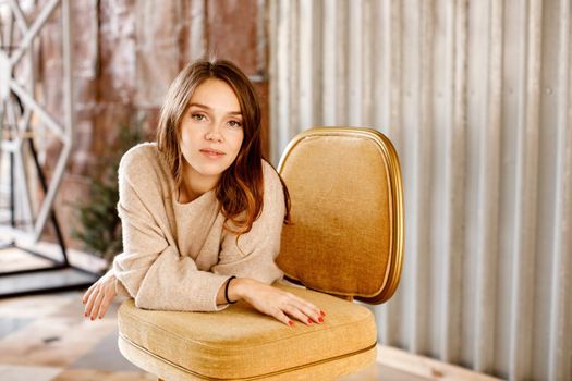 Charming female wearing cozy sweater leaning on chair and looking at camera while standing in contemporary room