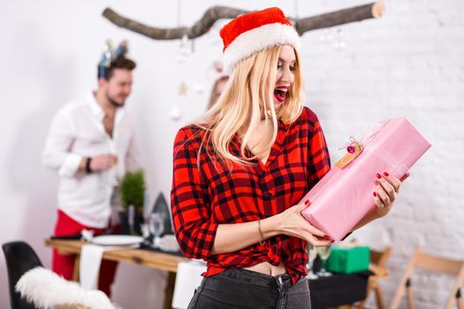 Portrait of a young woman with a pink giftbox on the foreground. Beautiful blonde in a Santa hat, red shirt in a cage and black jeans. Christmas party at home.
