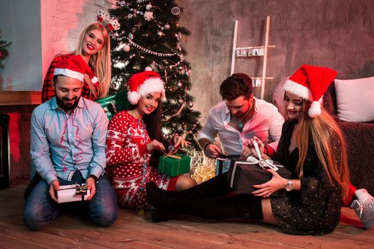Picture showing group of friends with Christmas presents on party at home. Young people sit on the floor near the Christmas tree and exchange gifts