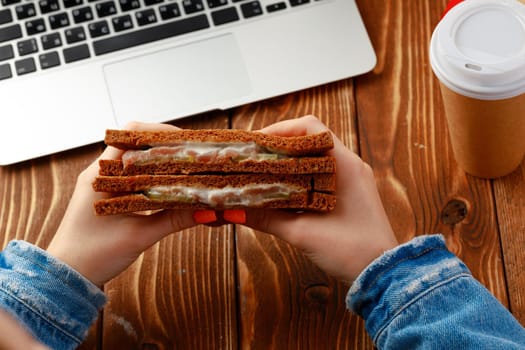 Hands of a woman holding sandwich above working table with open laptop