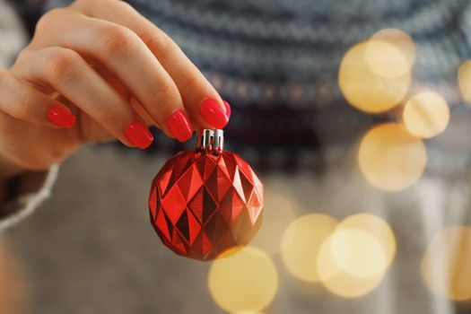 Hand of a woman in festive sweater holding Christmas bauble with blurred lights close up