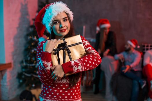 Portrait of a young woman with a gold giftbox on the foreground. Beautiful brunette in a Santa hat, red costume with deers. Christmas party at home.