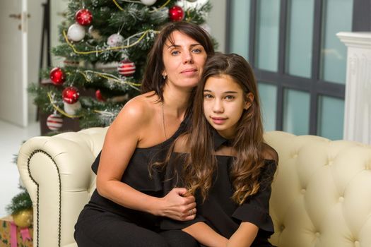 Happy mother and daughter near the Christmas tree are sitting on the couch. Family holidays concept.