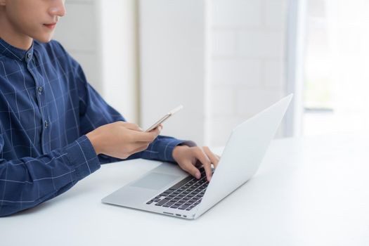 Closeup hand of young man working laptop computer and reading smartphone on internet online on desk at home, freelance male using phone with social media, business and communication concept.