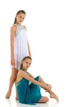 Two Beautiful Teenage Girls in White and Blue Sport Dresses Posing Together Against White Background, Portrait of Pretty Twin Sisters, One of Them Sitting on the Floor, Second Standing Behind