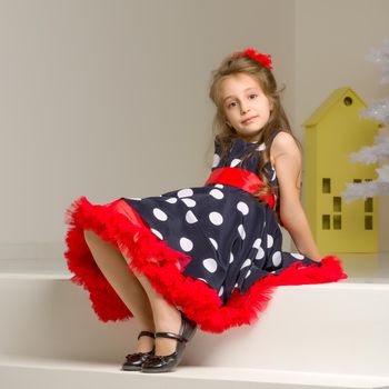 Portrait of Beautiful Girl Wearing Blue Polka Dot Dress, Red Bow and Black Shoes Sitting on White Stairs and Looking at Camera, Charming Coquettish Girl Posing in Retro Fashion Clothes in Studio