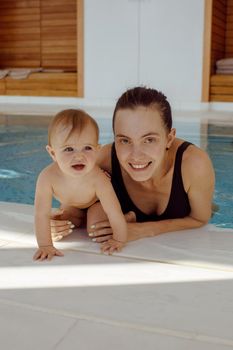 Cheerful young woman with adorable baby leaning on poolside while resting in clean water of swimming pool