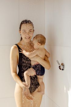 Smiling mother in bathing suit holding on hand baby standing under water in shower and looking at camera