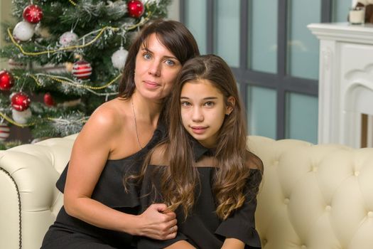 Happy mother and daughter near the Christmas tree are sitting on the couch. Family holidays concept.