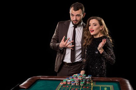 A man in a brown suit with a beautiful woman in a black dress is playing at a roulette table at a casino. Emotion of gambling players