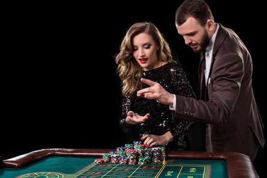 A man in a brown suit with a beautiful woman in a black dress is playing at a roulette table at a casino. Emotion of gambling players
