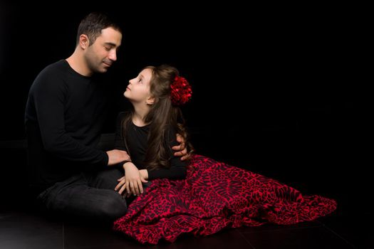 Portrait of Loving Father Hugging His Beautiful Daughter, Handsome Young Man Sitting on the Floor with his Adorable Girl, Happy Family Sitting Against Black Studio Background