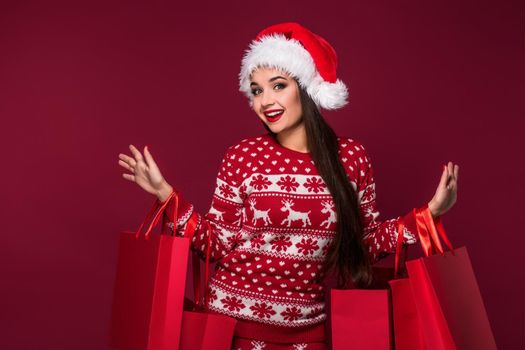 Young pretty woman or girl with long beautiful hair in Santa hat and New Year's dress holding red package on red studio background. Christmas sale. Emotional brunette. Copy space