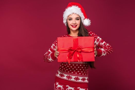 Young woman with long beautiful hair in Santa hat and New Year's dress holding Christmas present in her hands on red studio background. Christmas sale. Emotional brunette. Copy space