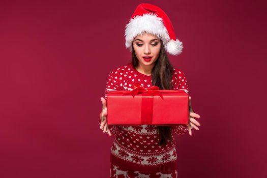 Young woman with long beautiful hair in Santa hat and New Year's dress holding Christmas present in her hands on red studio background. Christmas sale. Emotional brunette. Copy space