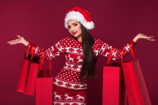 Young pretty woman or girl with long beautiful hair in Santa hat and New Year's dress holding red package on red studio background. Christmas sale. Emotional brunette. Copy space