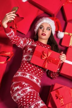 Funny young woman in a New Year's dress lying on a mountain of gifts. Christmas sale. Emotional brunette in Santa hat makes selfie