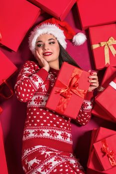 Funny young woman in a New Year's dress lying on a mountain of gifts. Christmas sale. Emotional brunette in Santa hat