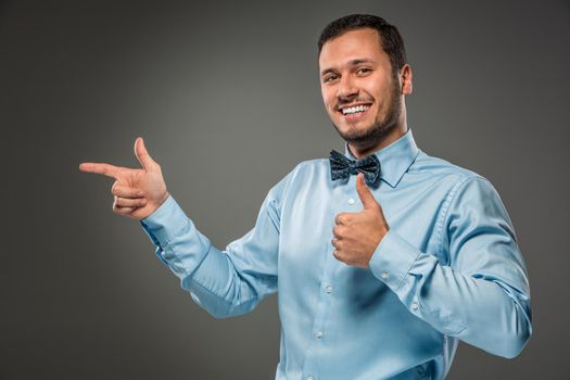 Closeup portrait of excited, energetic, happy, smiling man in blue shirt and butterfly tie is gesturing with his hand, pointing to something with an index finger, isolated on gray background. Positive human emotion facial expression