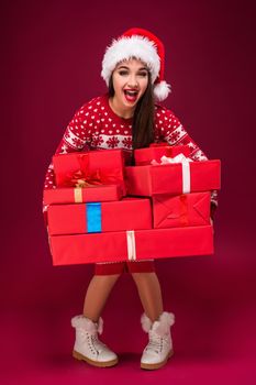 Amazed beautiful woman holding many presents on red background. Young woman in santa hat looks at camera. Christmas. Discounts. The shops. Sale